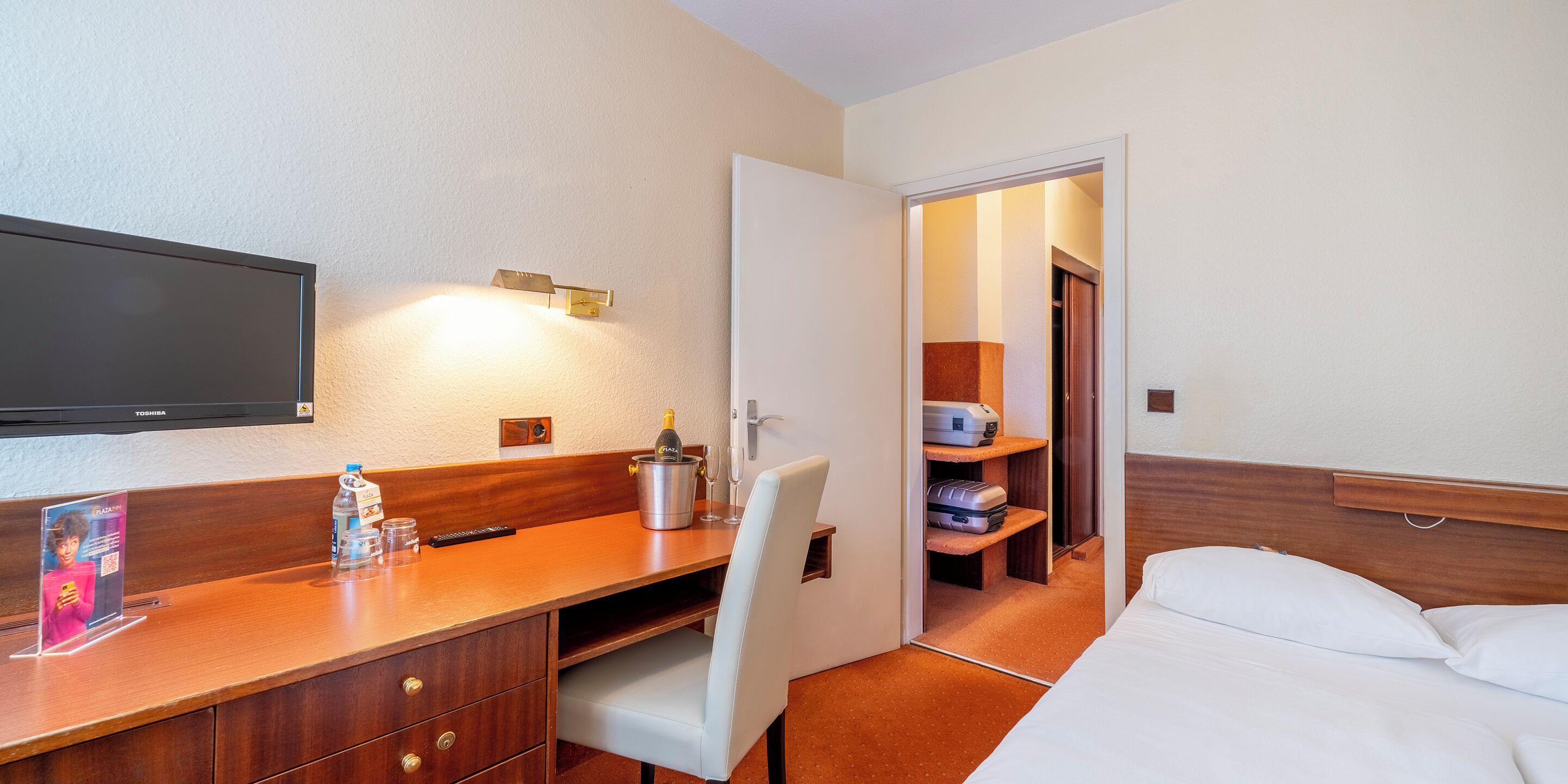 A hotel room with a wooden desk, a flat-screen TV, a minibar, a bed, and a walk-in closet.