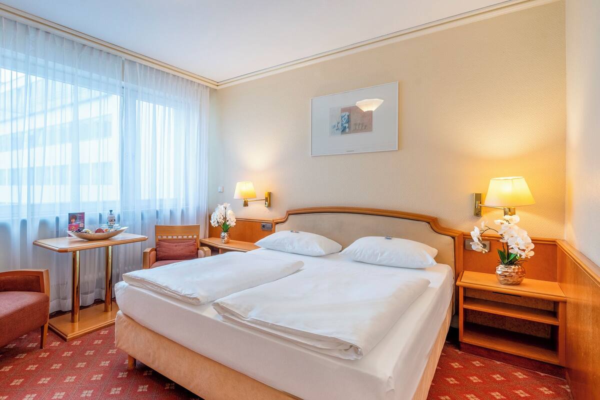 A hotel room with a double bed, a small table with a bowl and a bottle, a chair, a nightstand with a lamp, and a wall-mounted picture.