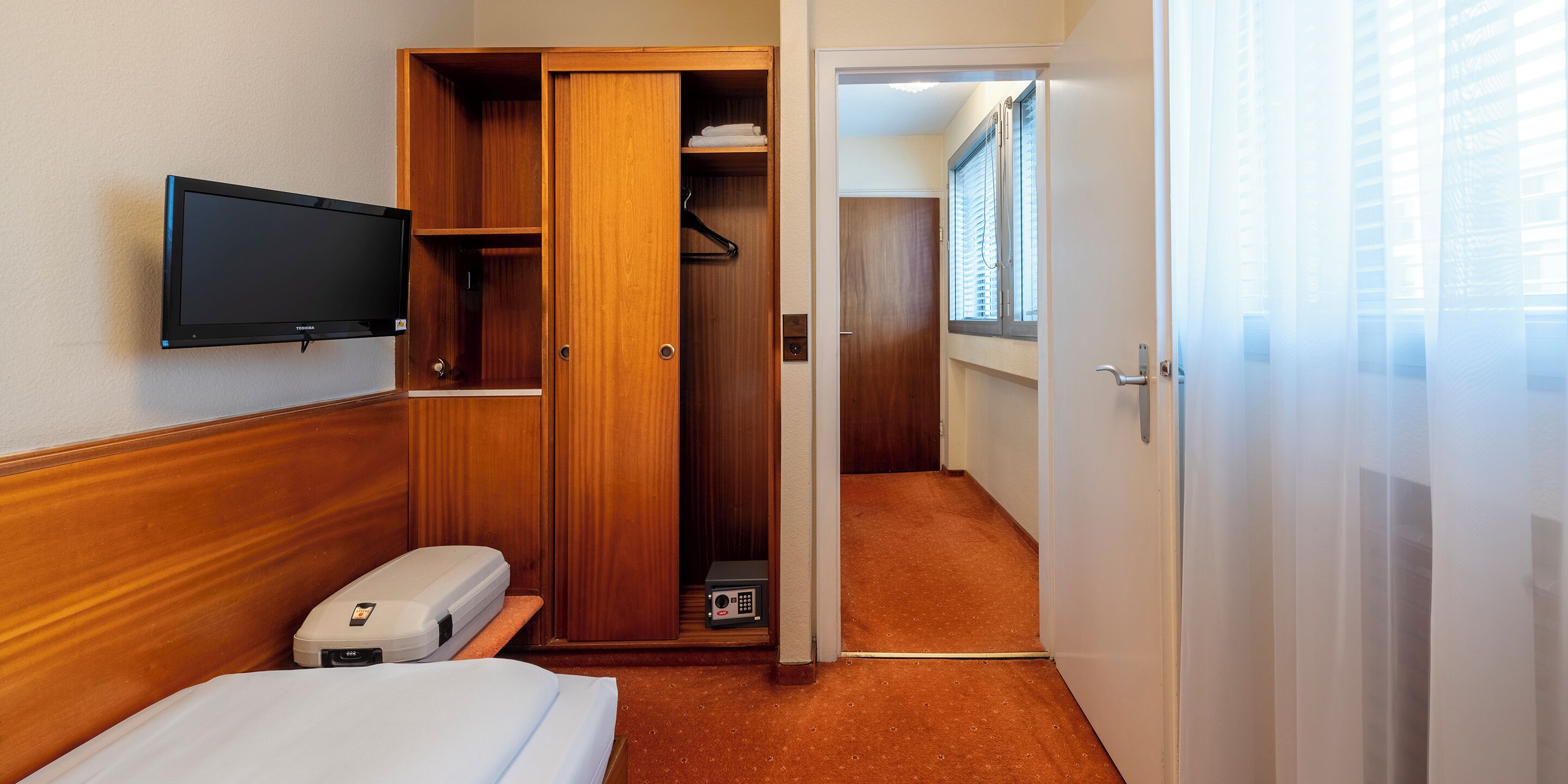 A hotel room with a bed, a TV, a wooden wardrobe, and a bathroom visible through an open door.