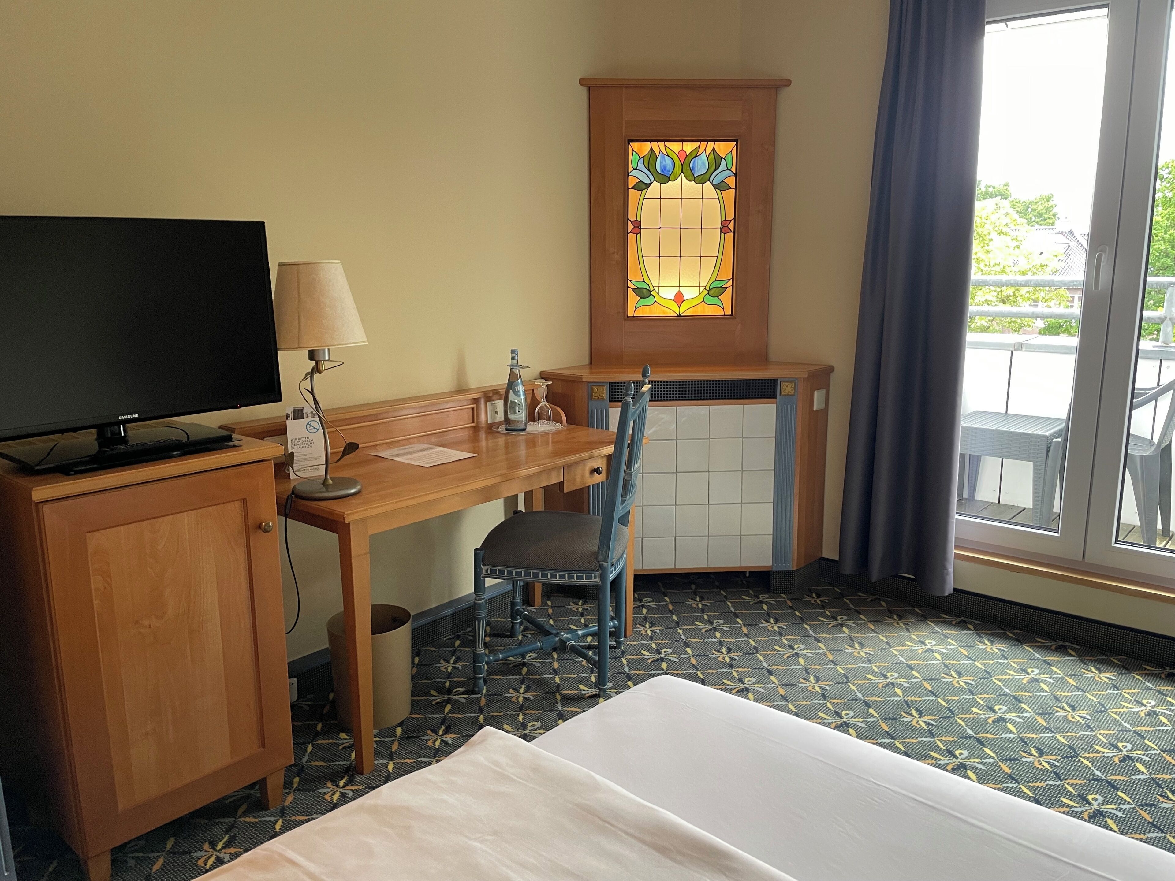 Une chambre d’hôtel comprenant un lit, un bureau avec une télévision, une chaise, une petite table, une fenêtre avec vue et un vitrail décoratif.