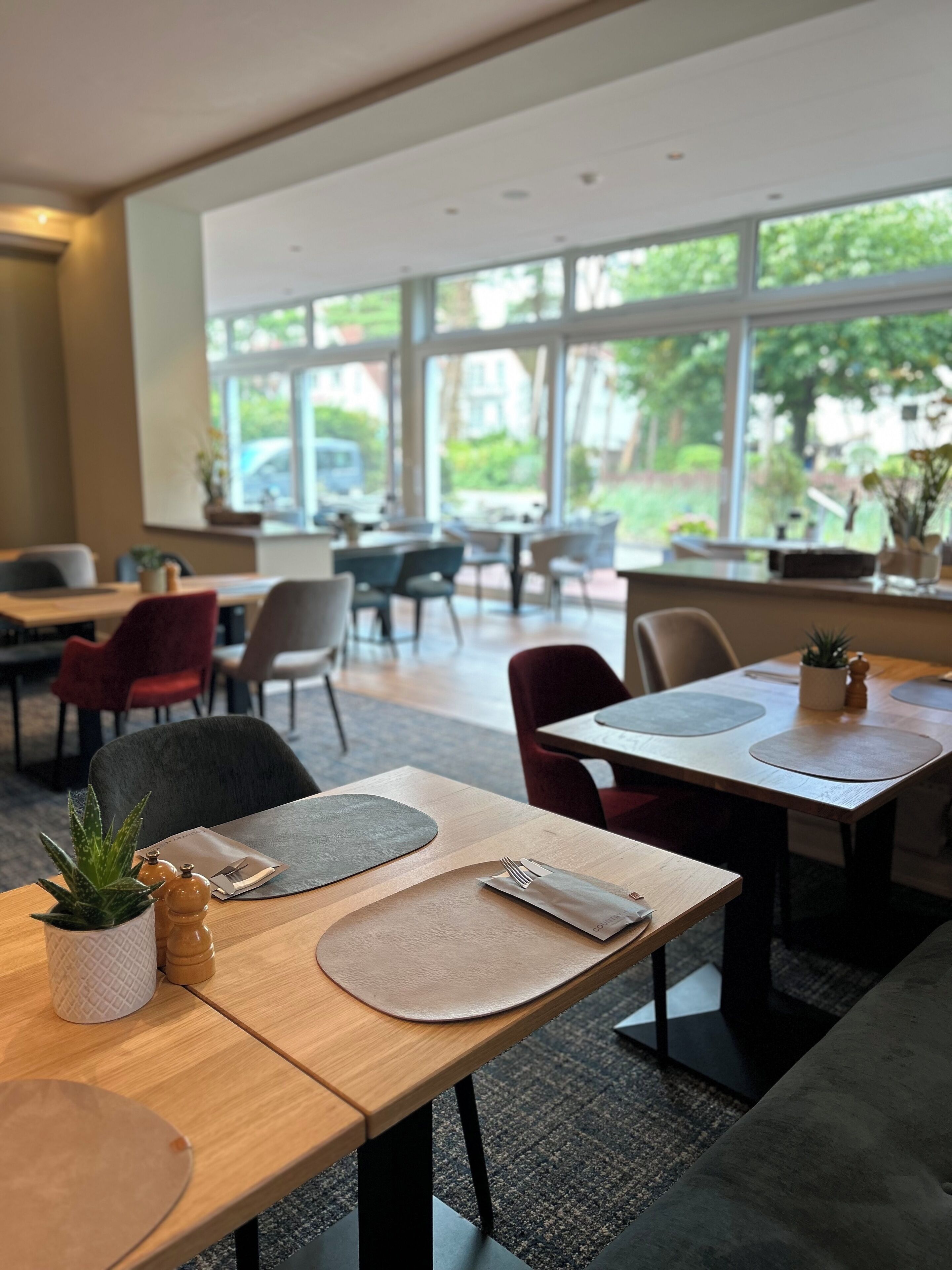 A modern dining area with tables set for two, featuring minimalist decor and large windows offering a view of the outdoors.