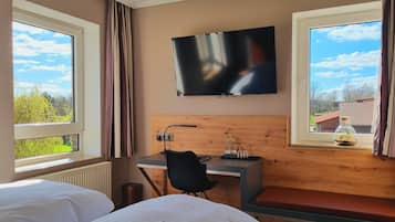 Een hotelkamer met twee bedden, een bureau, een televisie en uitzicht op groen en gebouwen.