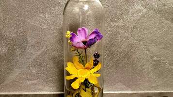 Eine klare Glasflasche mit Korkverschluss, gefüllt mit gelben und violetten Blumen, vor einer strukturierten Wand platziert.