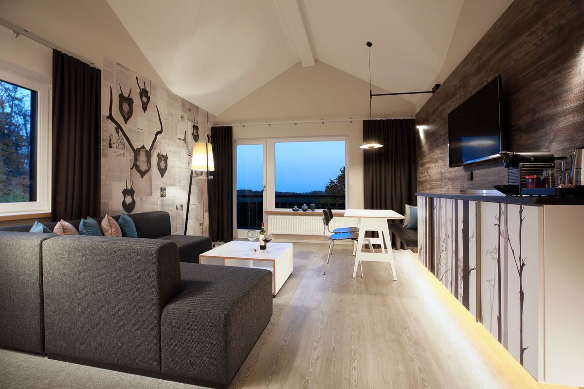 A modern living room with a sectional sofa, a coffee table, a dining area with a table and chairs, a flat-screen TV mounted on a wooden wall, and a large window with a view of trees.