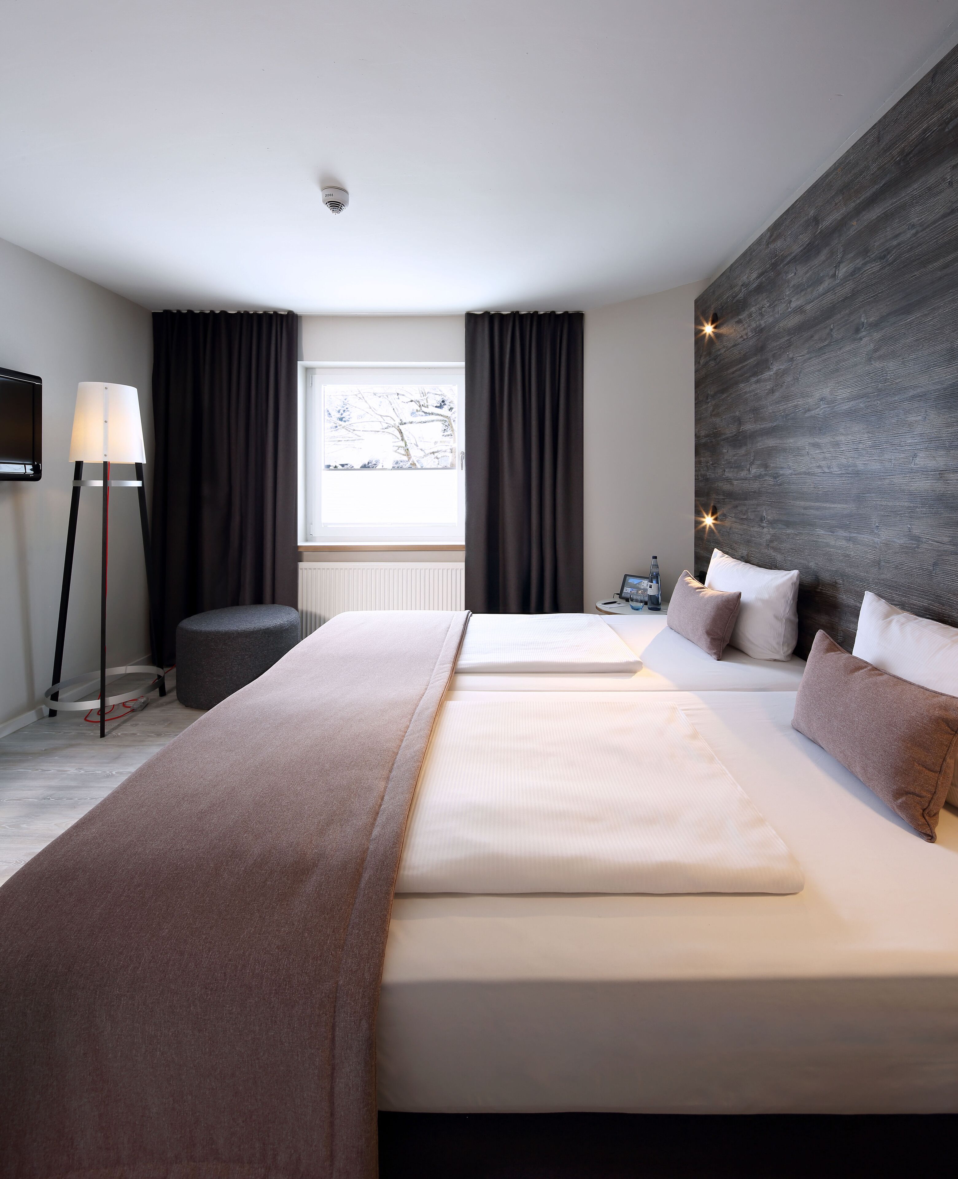 A modern hotel room with two beds, a large window with curtains, a floor lamp, and a view of trees outside.