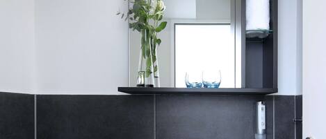 Un baño moderno con un lavabo blanco, baldosas oscuras, un estante de vidrio con un jarrón de flores y un inodoro.