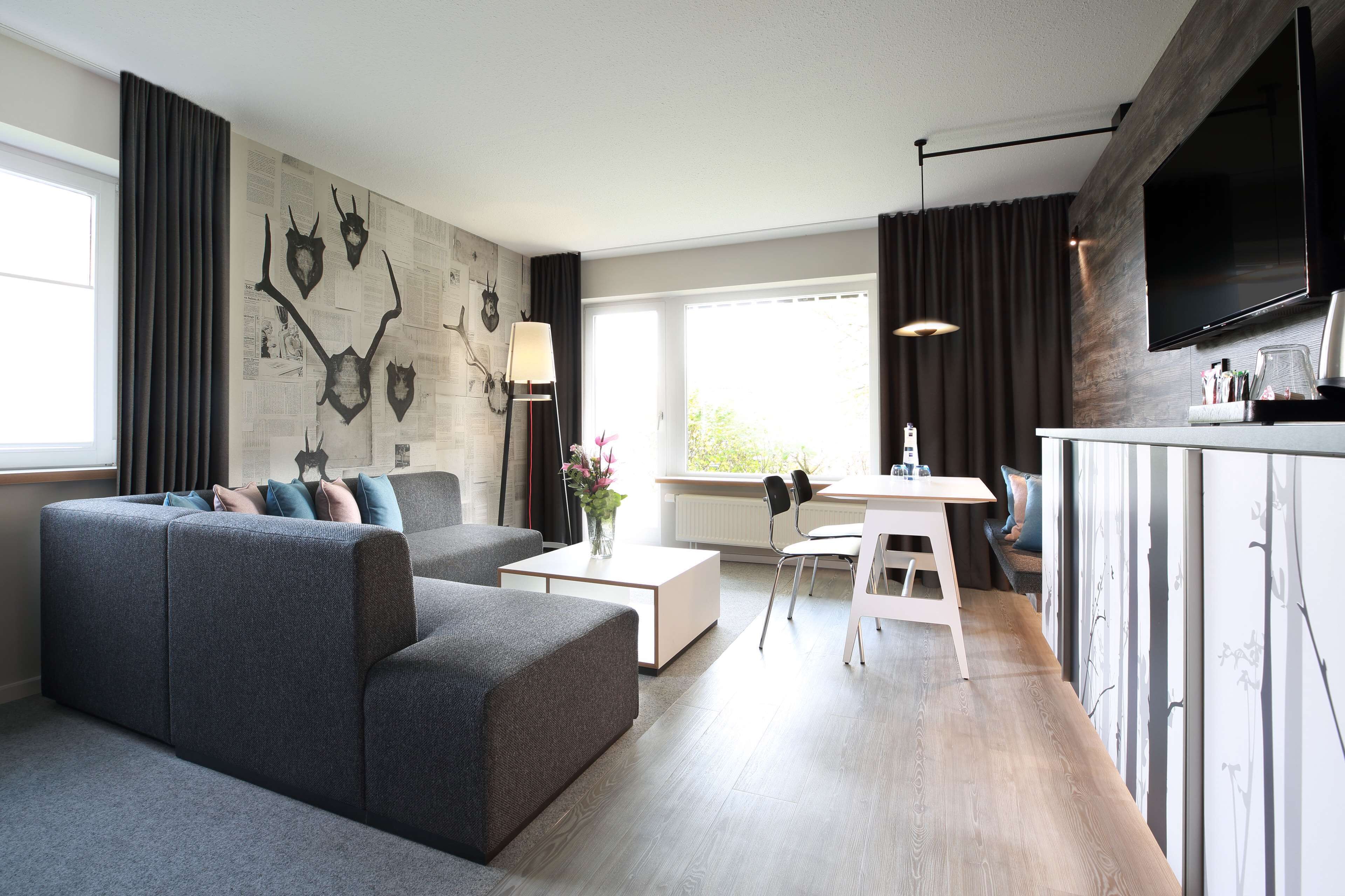 A modern living room with a grey sectional sofa, a white coffee table, and a dining area with a white table and chairs. A large flat-screen TV is mounted on the wall, and there is a window with curtains.
