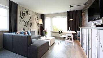 Una sala de estar moderna con un sofá seccional gris, una mesa de centro blanca y un comedor con una mesa y sillas blancas. Un televisor de pantalla plana grande está montado en la pared, y hay una ventana con cortinas.