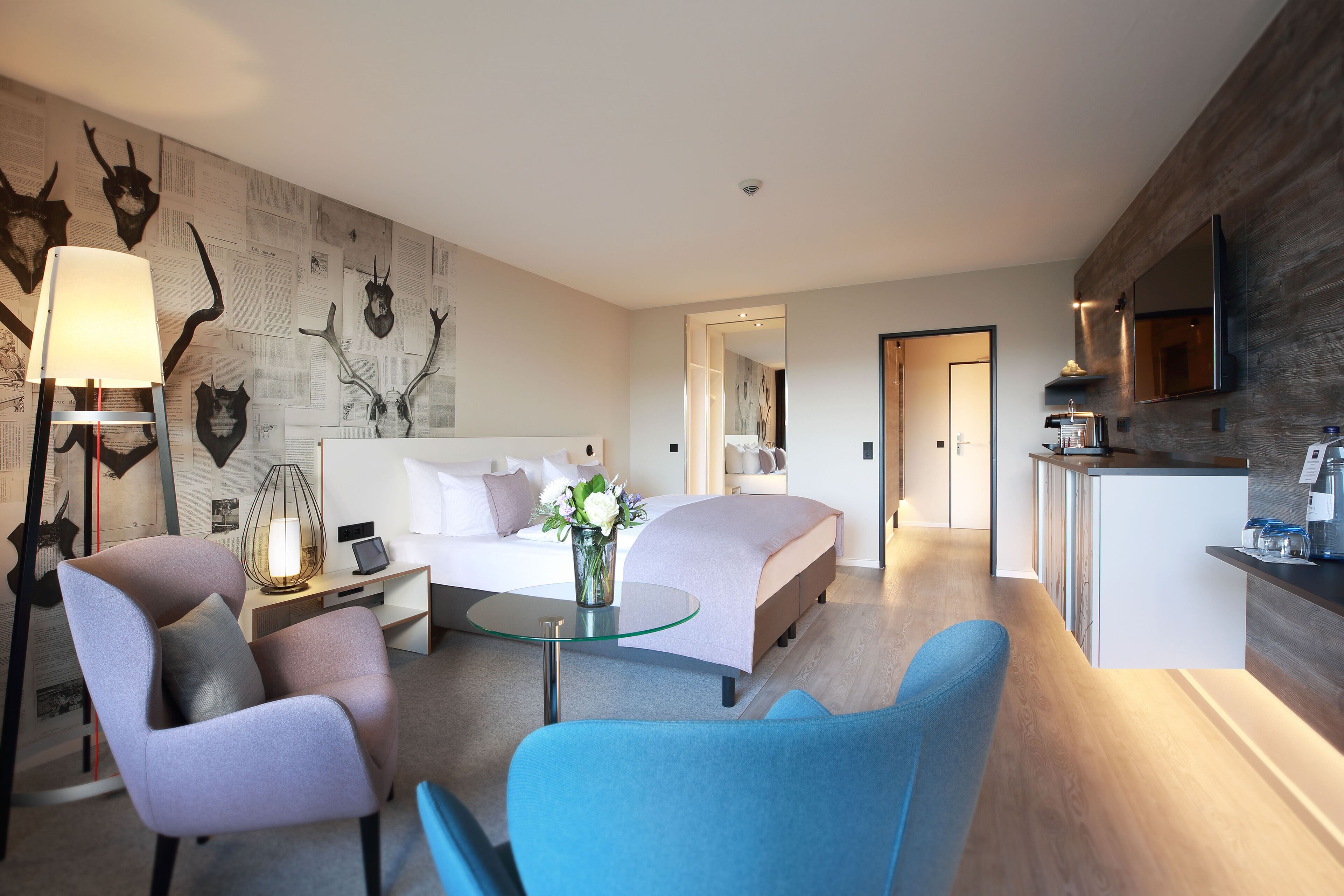 A modern hotel room with a bed, a desk, a chair, a glass coffee table, and a wall with mounted animal heads.