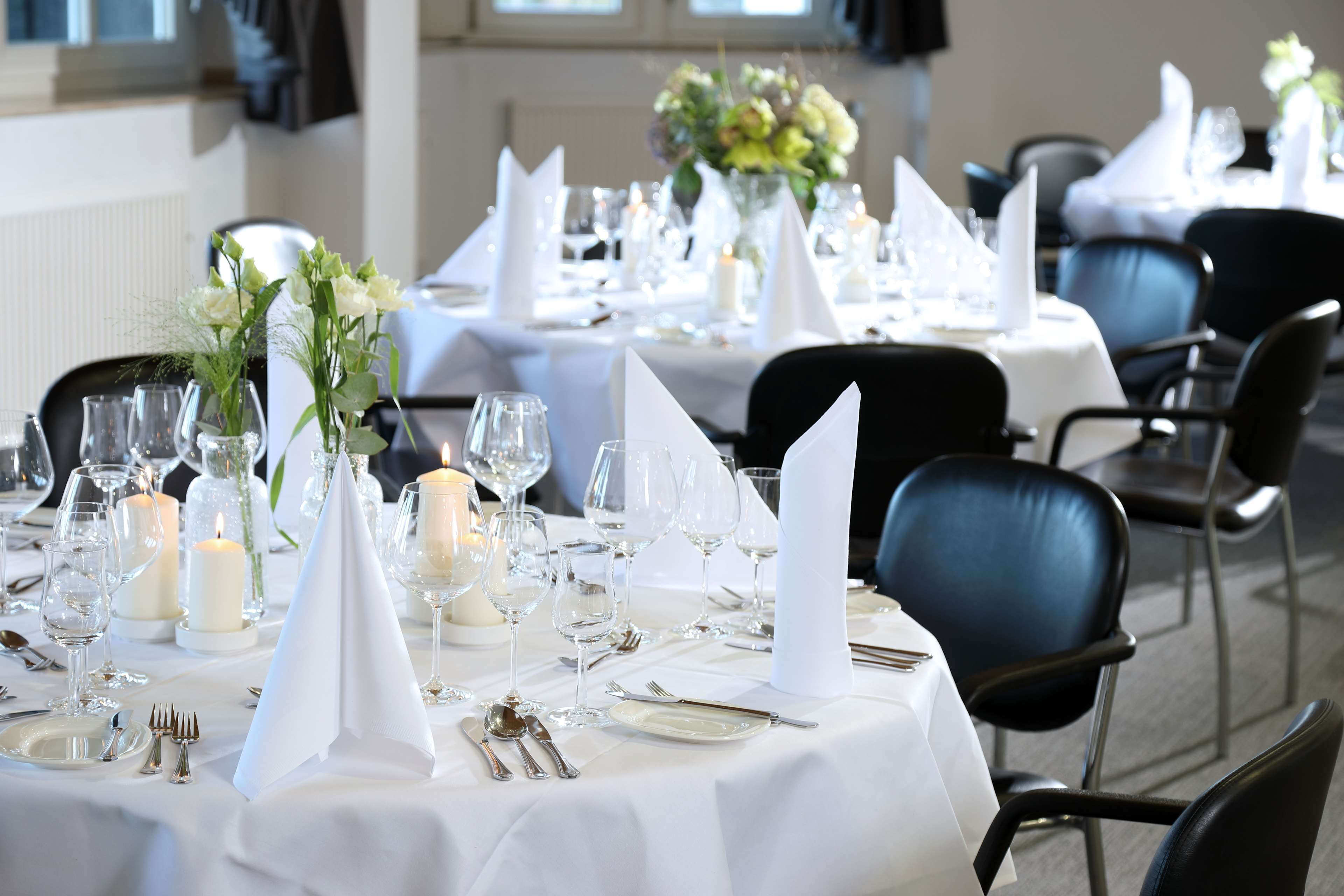 A dining area with tables set for a formal event, featuring white tablecloths, candles, and floral centerpieces.