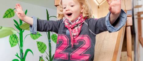 Un niño en un tobogán de madera con el número 22 en su camiseta, rodeado de una zona de juegos con una casita de madera y un mural pintado en la pared.