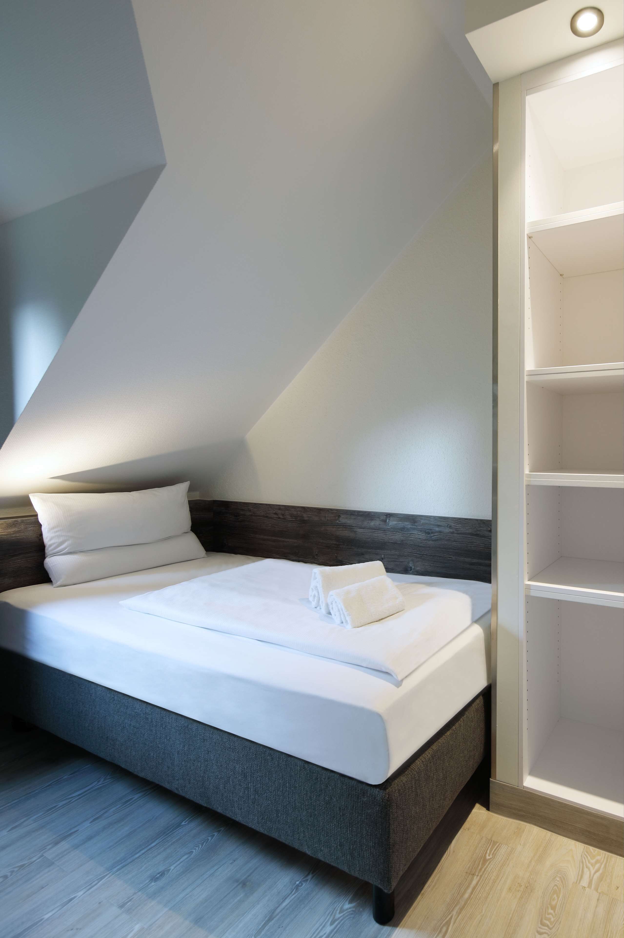 Una cama bien tendida en una habitación con techo inclinado, cabecera de madera y estantería empotrada.