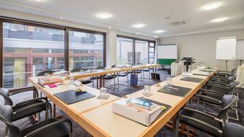 Ein Konferenzraum mit langen Tischen, Stühlen, einer Whiteboard-Tafel und Blick auf Gebäude durch große Fenster.