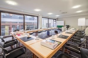 Un salón de conferencias con mesas largas, sillas, una pizarra y vistas a edificios a través de amplias ventanas.