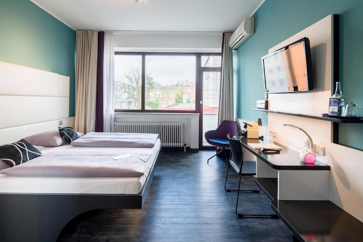 Ein Hotelzimmer mit zwei Betten, einem Schreibtisch, einem Sessel, einem Fernseher und einem Fenster mit Blick auf Gebäude.
