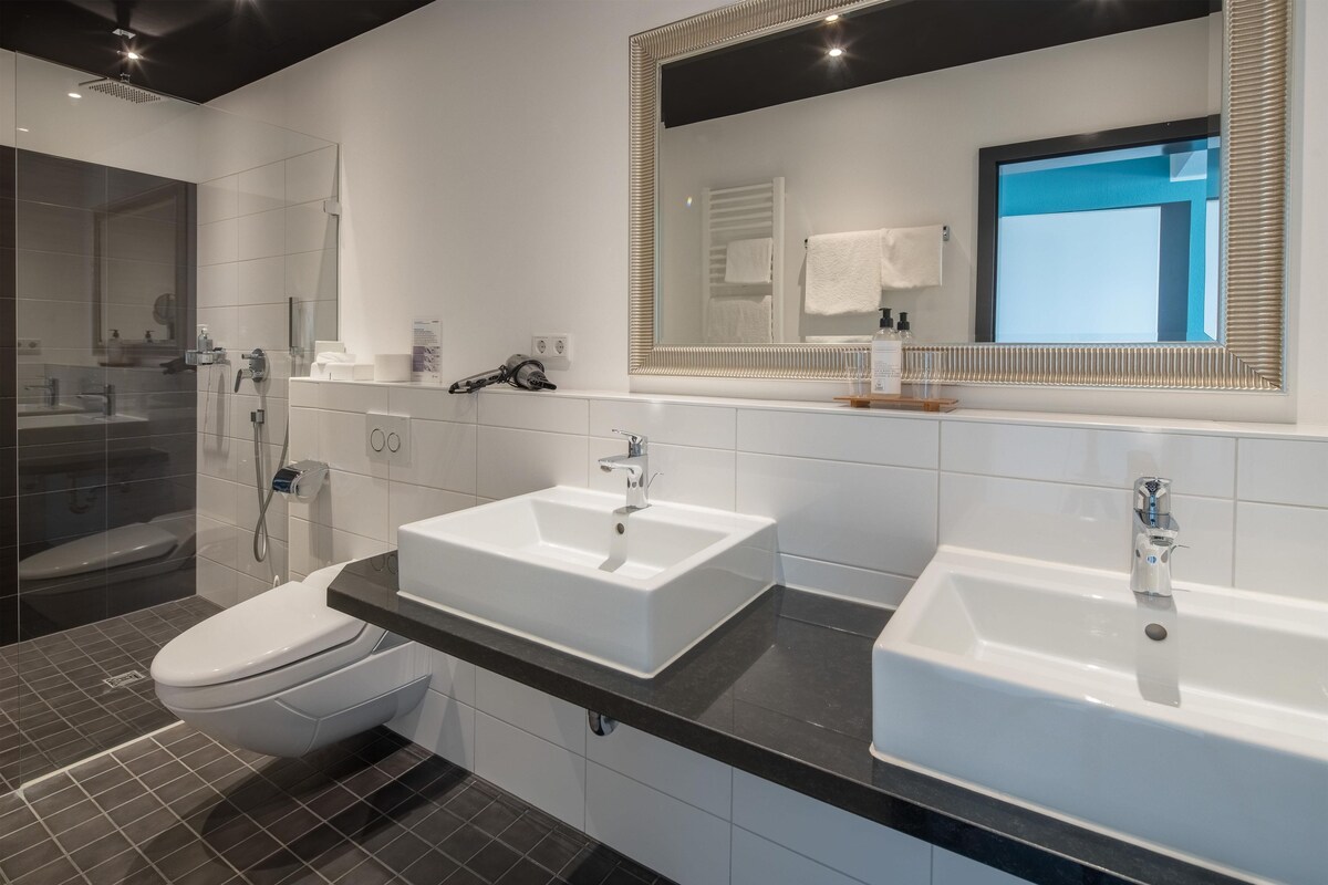Un baño moderno con ducha, dos lavamanos, inodoro y un espejo grande.