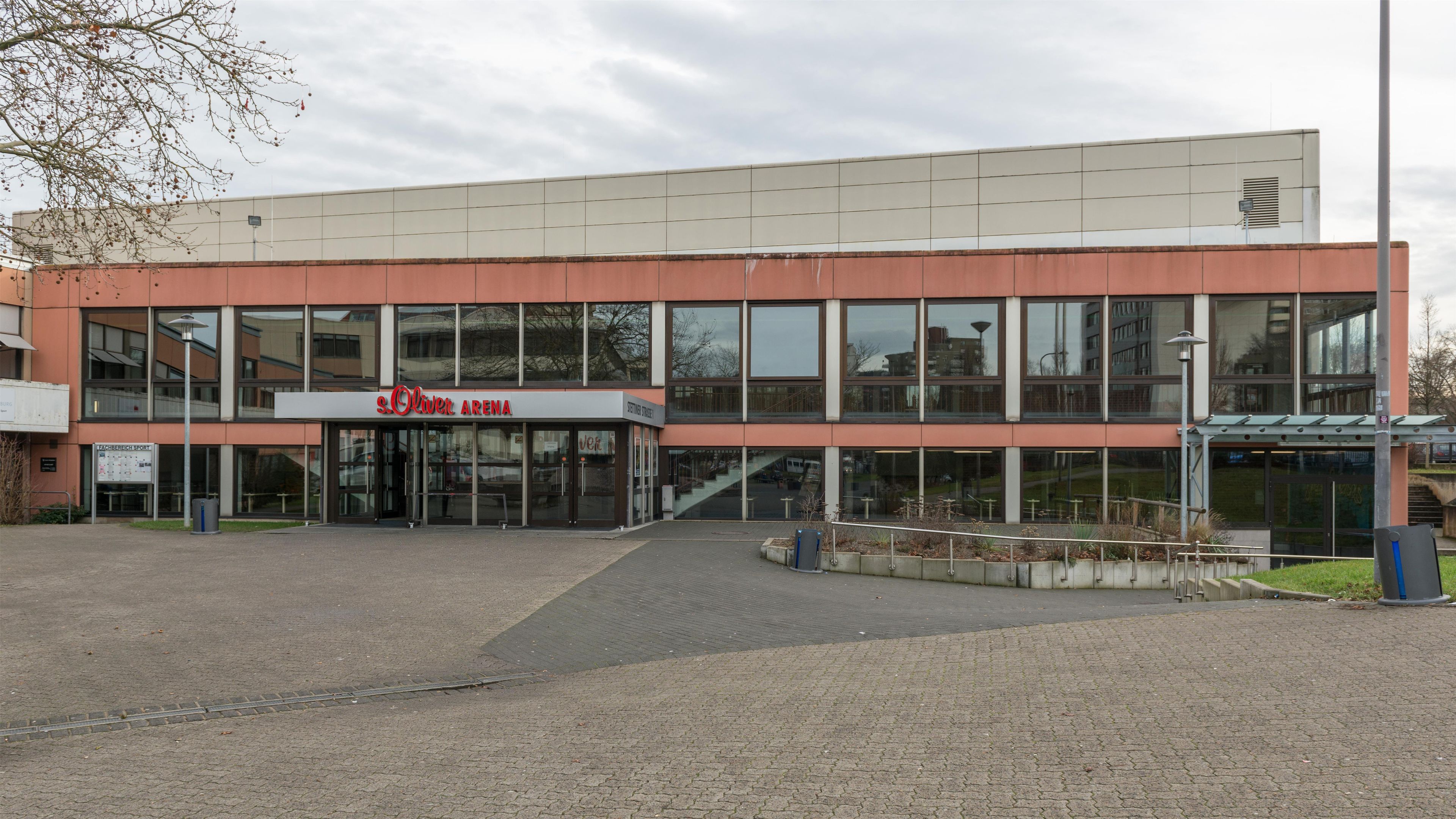 Un edificio moderno con un cartello che indica 'Oliver Arena'. Presenta una facciata in vetro, un ingresso coperto e un'area pavimentata antistante.