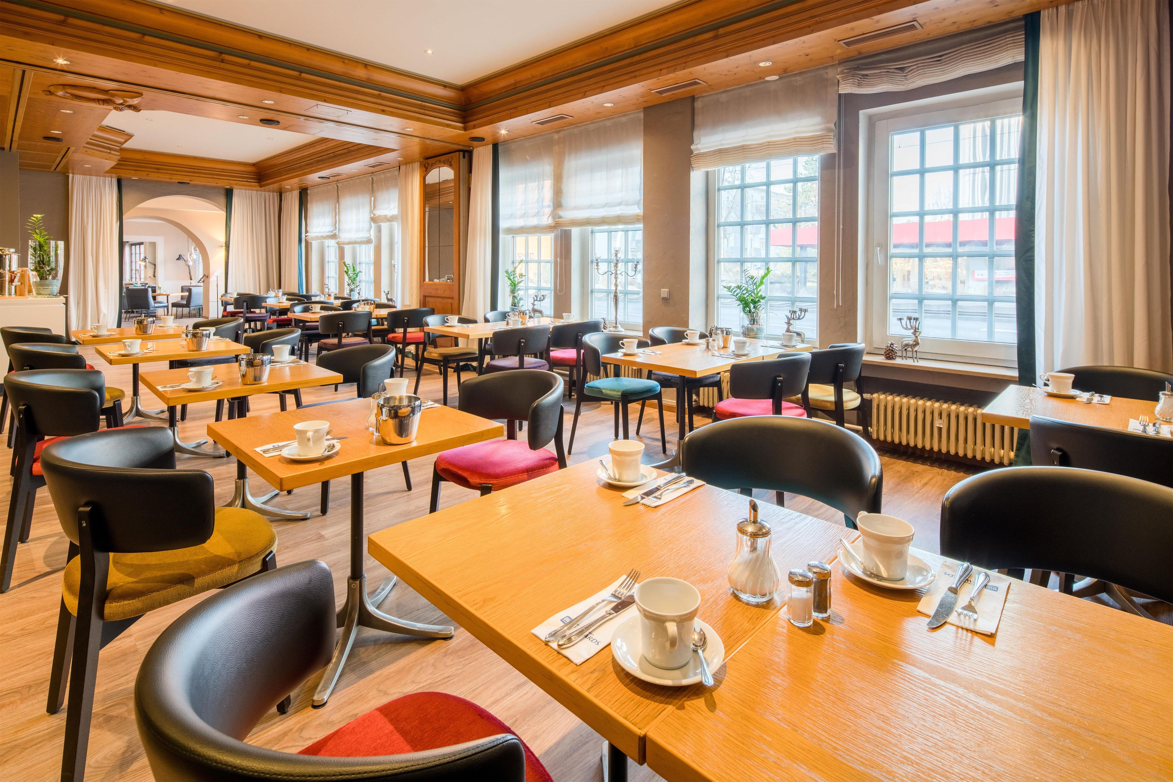 A hotel dining area with multiple tables and chairs, large windows, and a view of the street.
