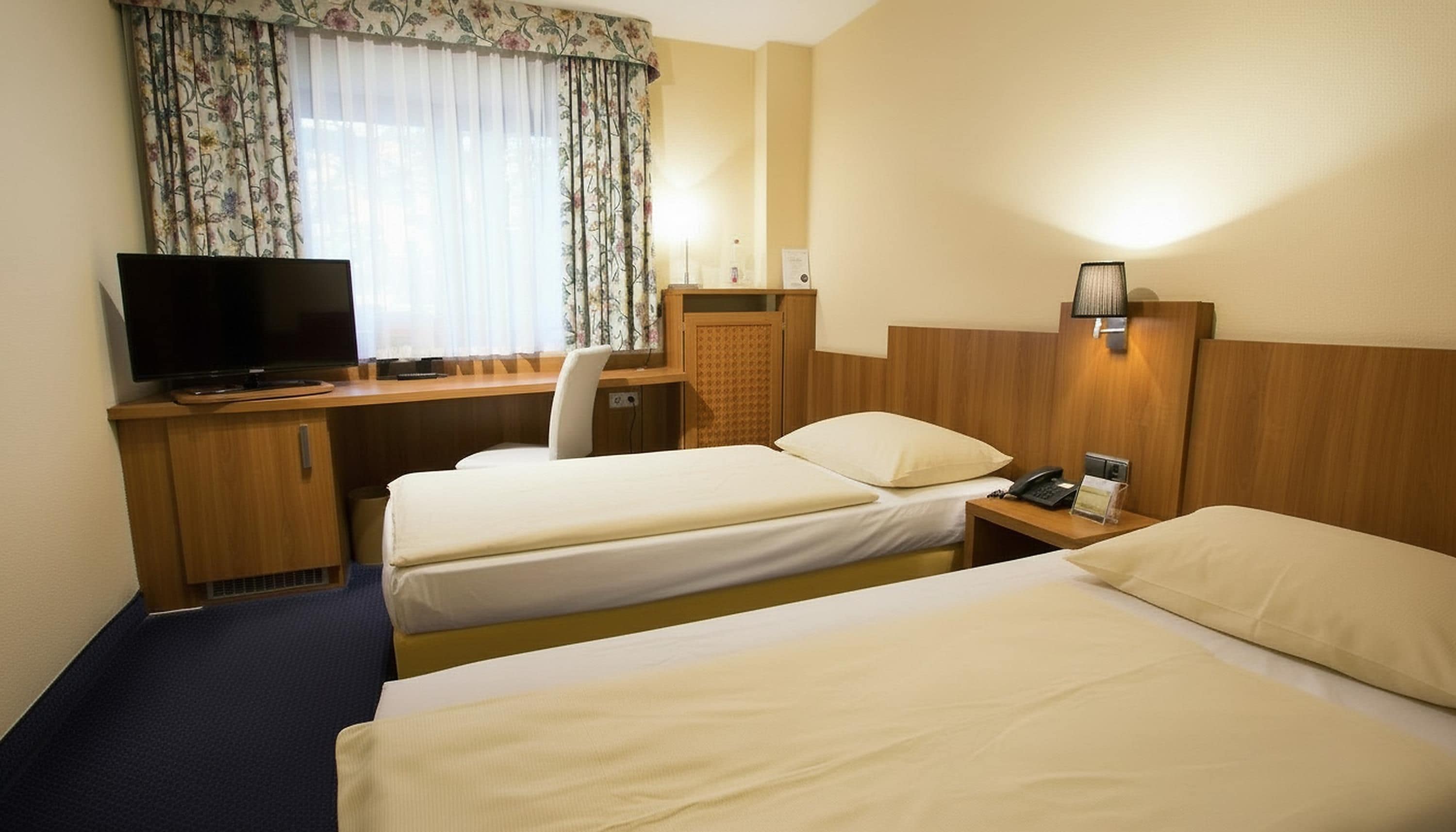 A hotel room with two beds, a desk with a television, a lamp, and a window with curtains.