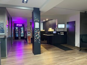 Eine Hotelhalle mit einer Rezeption, einem Self-Check-in-Terminal und einem Barbereich.