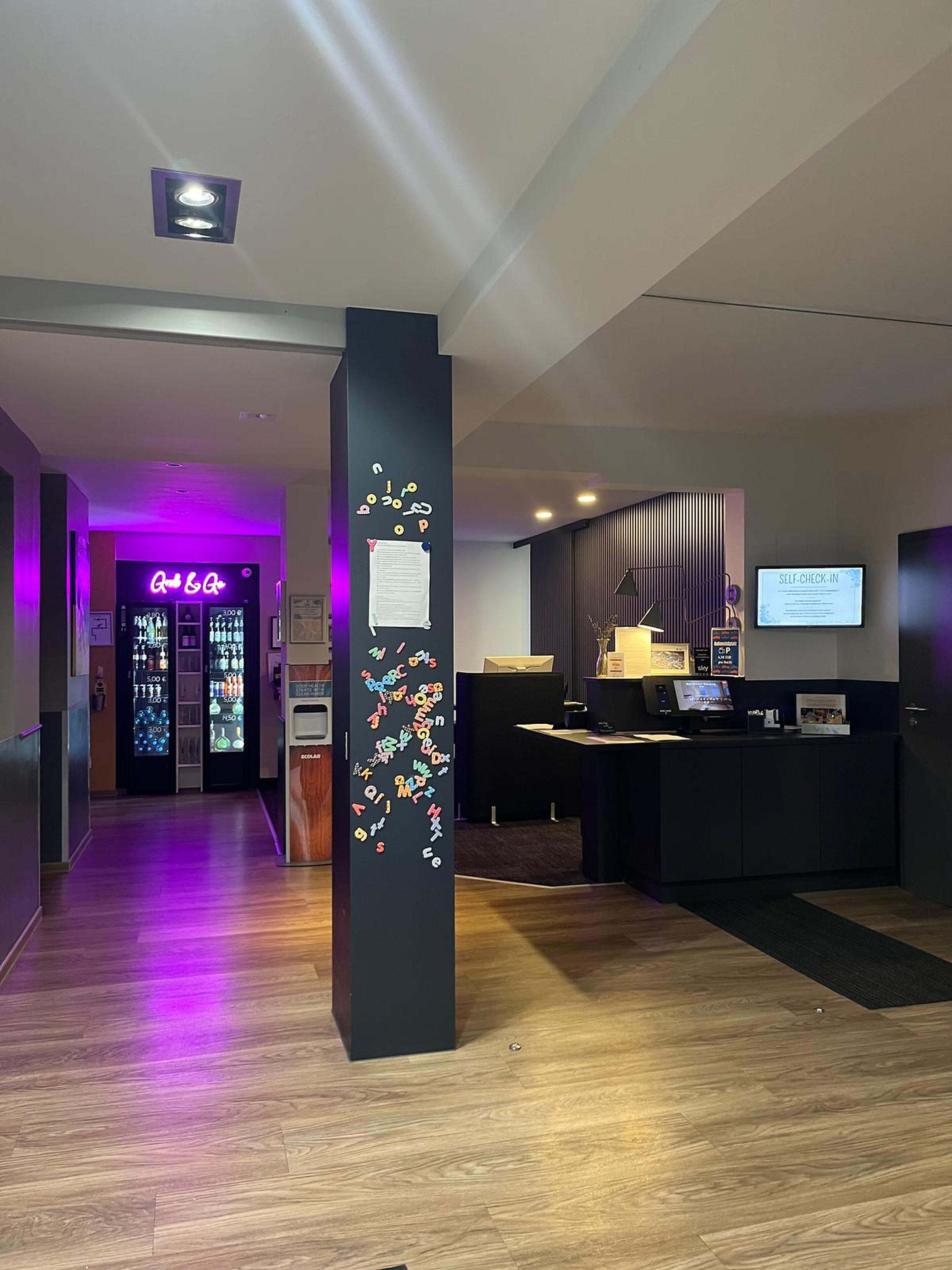 A hotel reception area with a reception desk, a self-check-in kiosk, and a neon sign in the background.