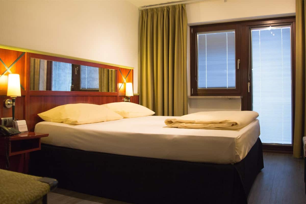 Ein Hotelzimmer mit Bett, Nachttischlampen, einem Spiegel, einem Fenster mit Vorhängen und einem Schreibtisch mit Telefon.