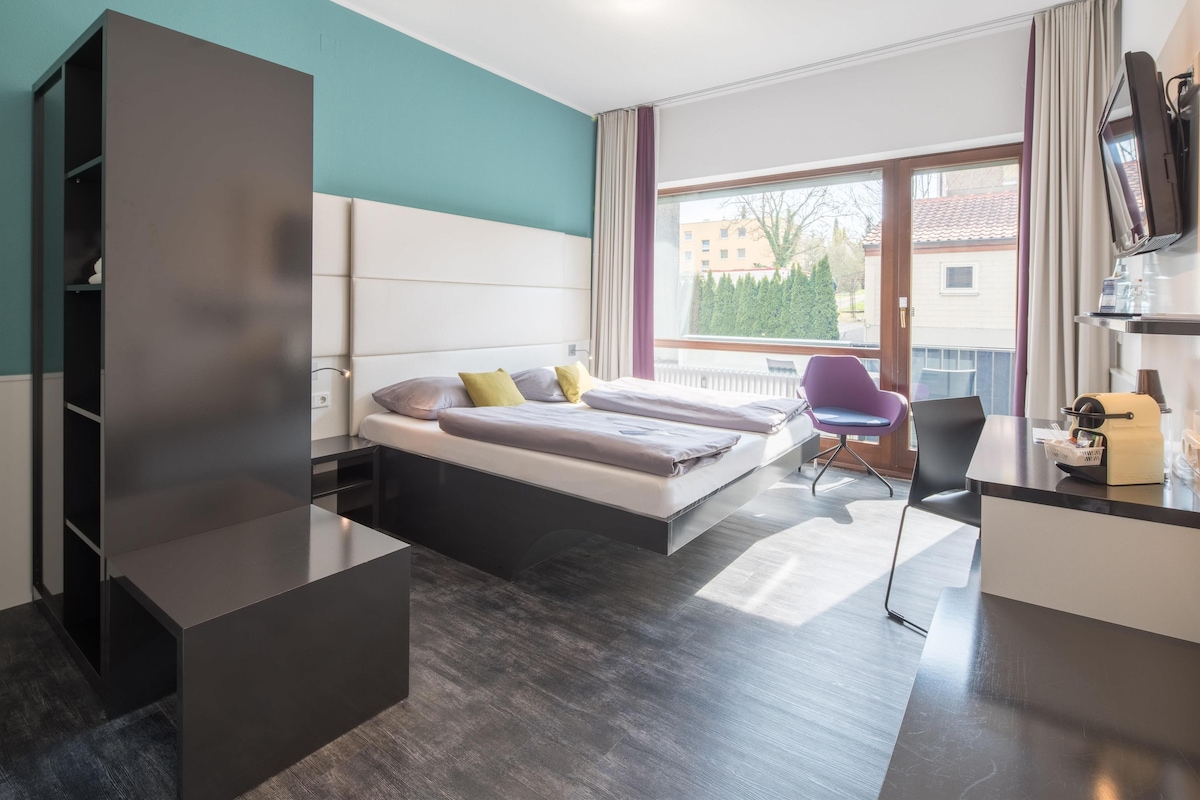 Quarto de hotel moderno com cama, escrivaninha, cadeira e televisão.