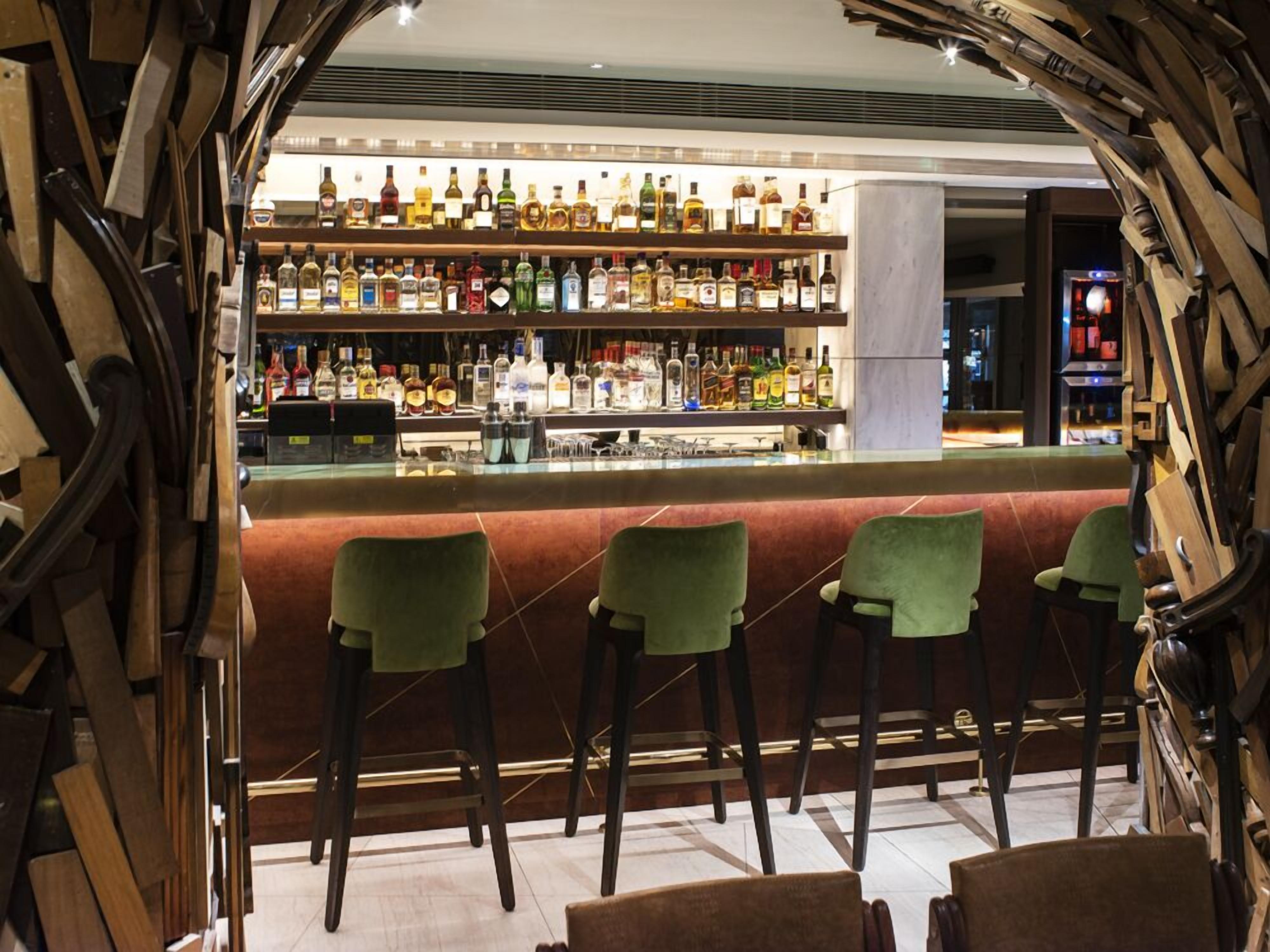 Un espace bar doté de nombreuses bouteilles d’alcool, de tabourets de bar verts et d’un design moderne agrémenté d’éléments en bois.