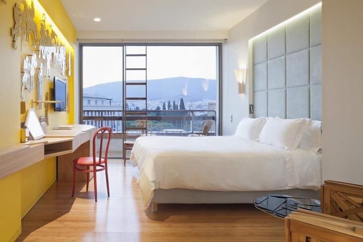 Habitación de hotel con cama, escritorio, silla y vista a un paisaje urbano a través de un ventanal grande.