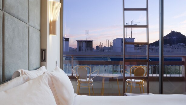 A hotel room with a large bed, two chairs, a balcony with a view of the city and a mountain. - New Hotel (Athens)