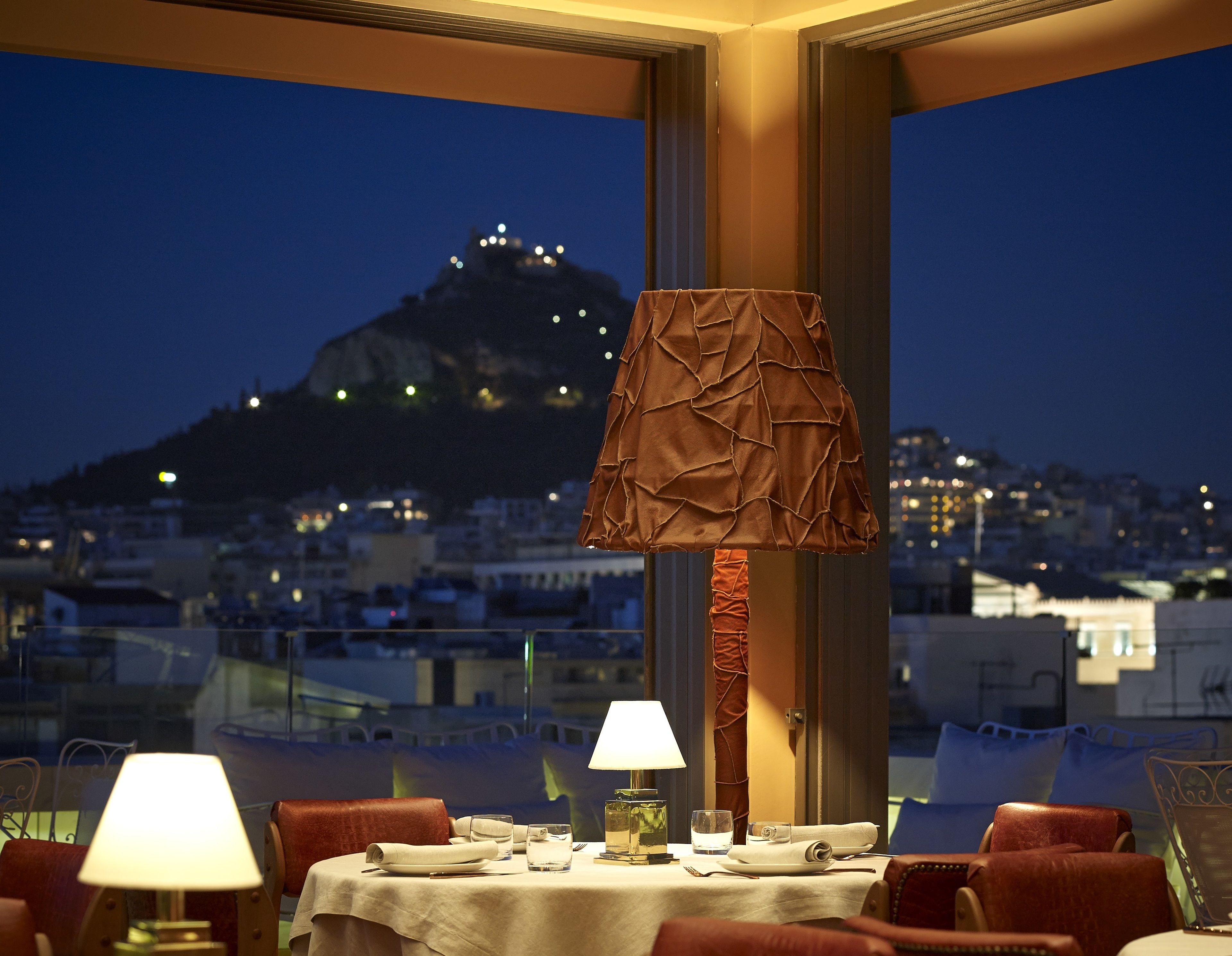 A dining table set with a lamp, glasses, and a bottle, overlooking a cityscape with a mountain in the background.