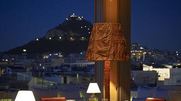 A dining table set with a lamp, glasses, and a bottle, overlooking a cityscape with a mountain in the background.