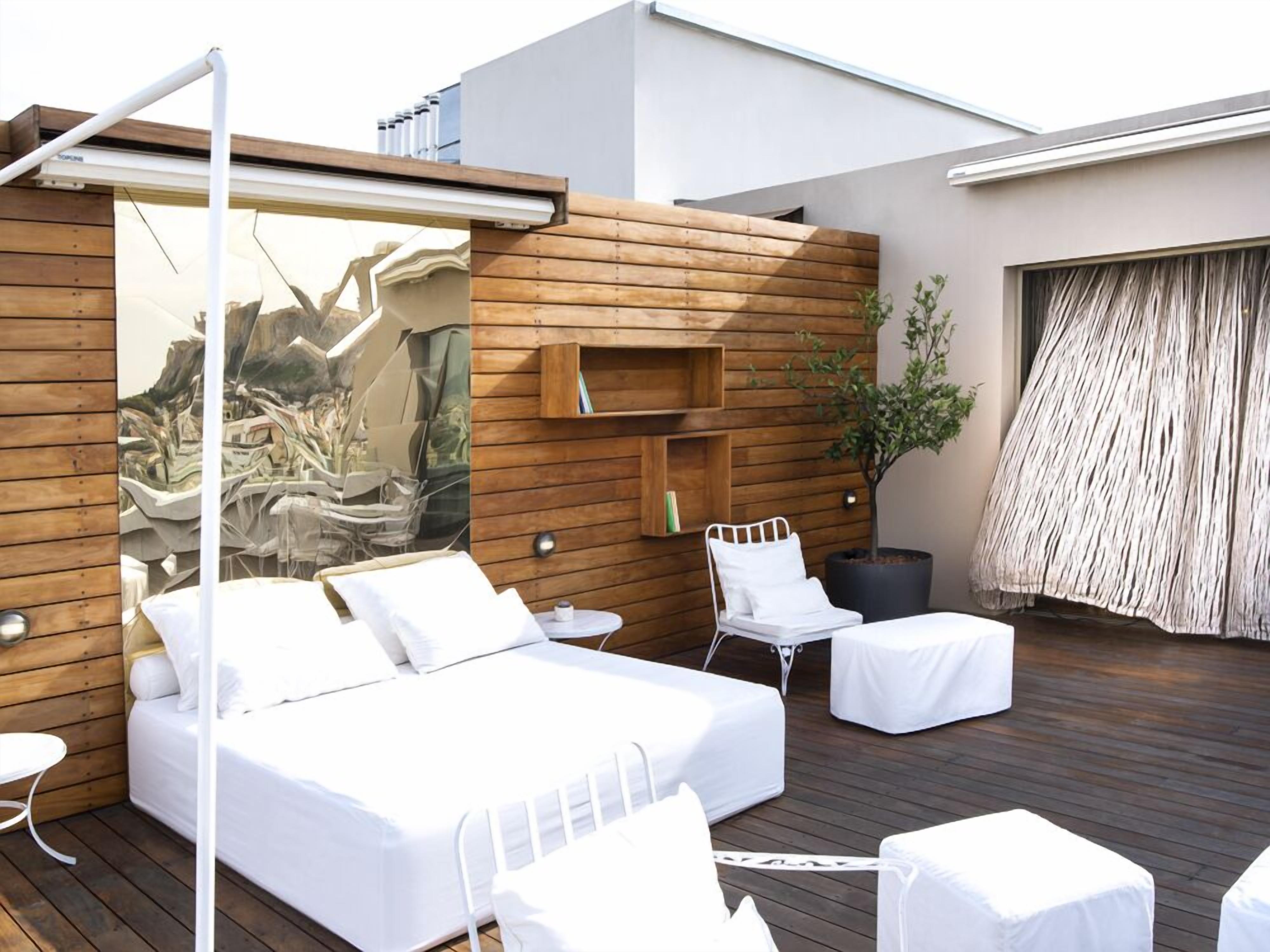 A modern outdoor bedroom with a bed, two white chairs, a small table, and a wooden wall with built-in shelves.