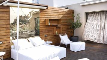 A modern outdoor bedroom with a bed, two white chairs, a small table, and a wooden wall with built-in shelves.