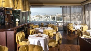 A restaurant with round tables and chairs, a bar area, and a view of the cityscape. - New Hotel (Athens)