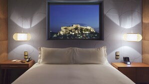A hotel room with a large bed, two bedside tables, a nightstand, a wall-mounted TV, and two wall-mounted lamps. - New Hotel (Athens)