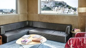 A modern room with a leather sofa, a round coffee table with books, and large windows offering a view of a historic cityscape. - New Hotel (Athens)