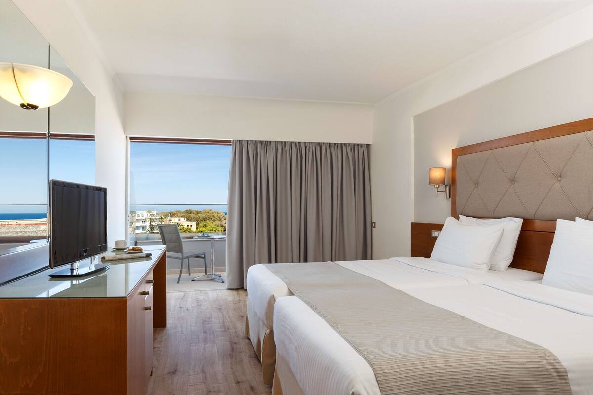 A hotel room with a bed, a desk with a TV, a chair, and a view of the sea.