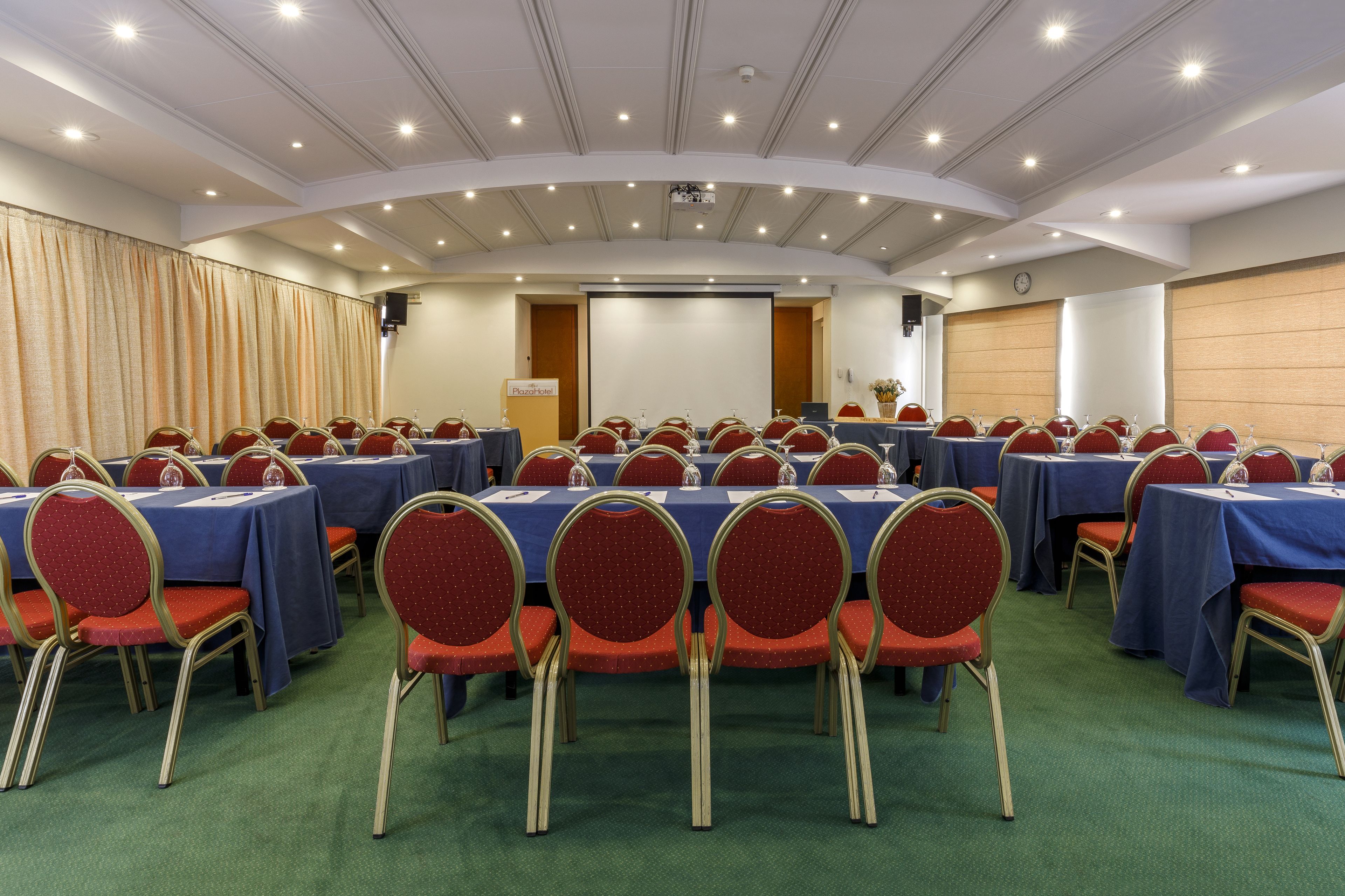 A conference room with multiple tables and chairs, a projector screen, and a clock on the wall.