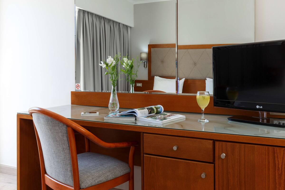 A hotel room with a wooden desk, a chair, a flat-screen TV, a vase with flowers, a magazine, and a glass of wine.