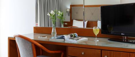 A hotel room with a wooden desk, a chair, a flat-screen TV, a vase with flowers, a magazine, and a glass of wine.