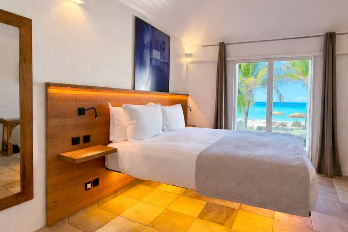 A hotel room with a bed, a wooden headboard, a nightstand, a large window with a view of the beach, and a ceiling with wooden beams.