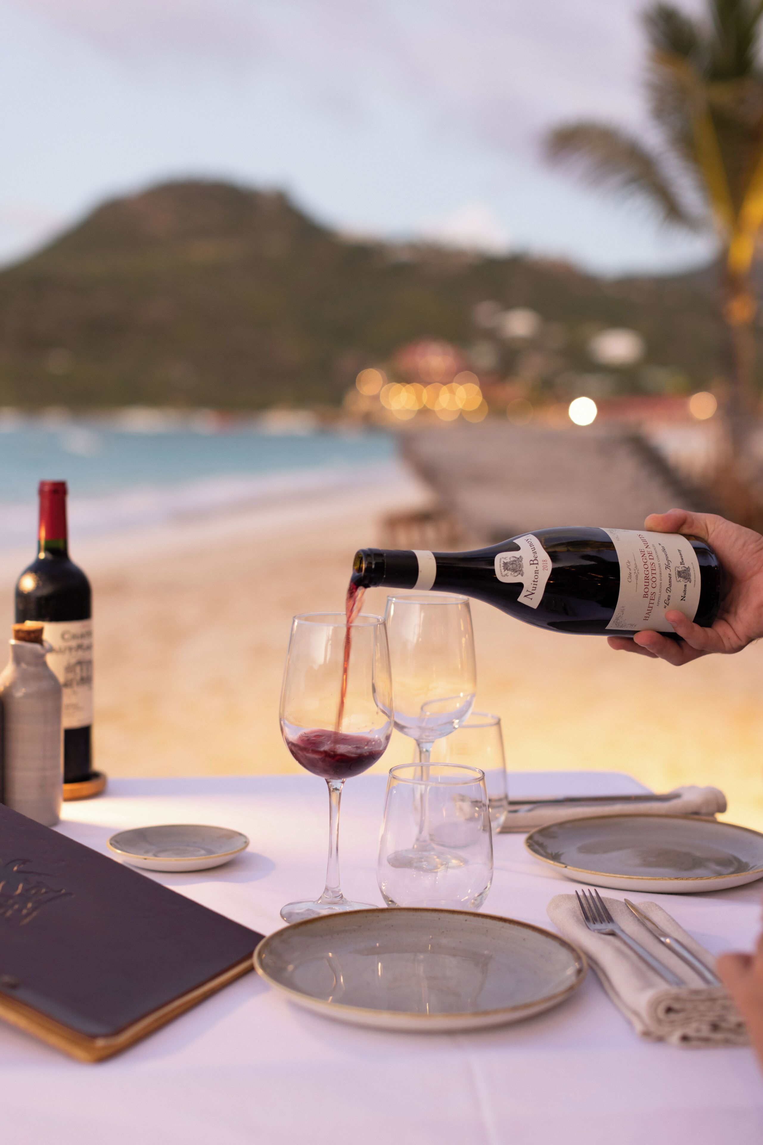 Una mesa para dos, con copas de vino, una botella y un menú, en una playa con una montaña de fondo.