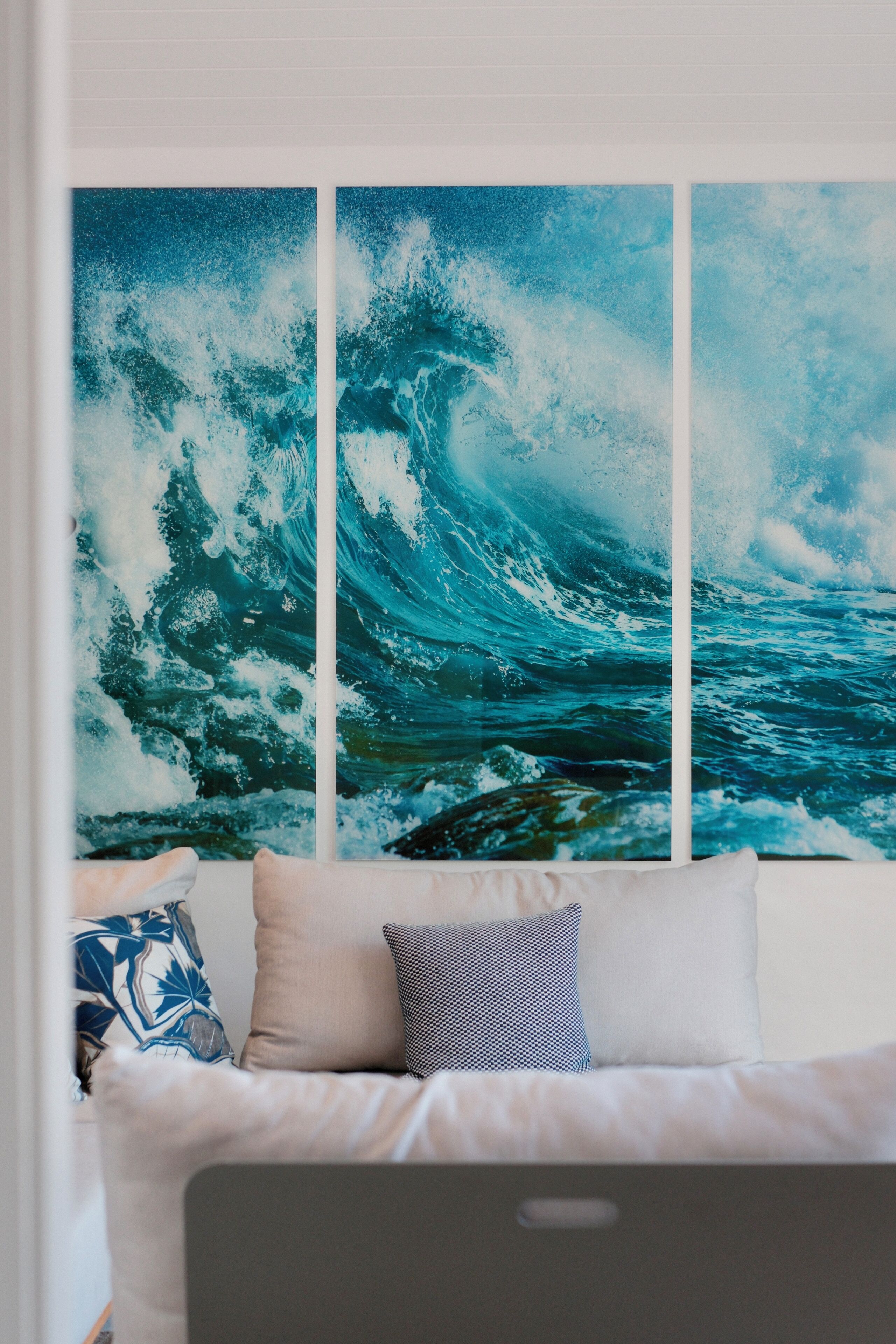 A room with a large ocean wave painting on the wall, a white sofa with patterned cushions, and a laptop on the sofa.