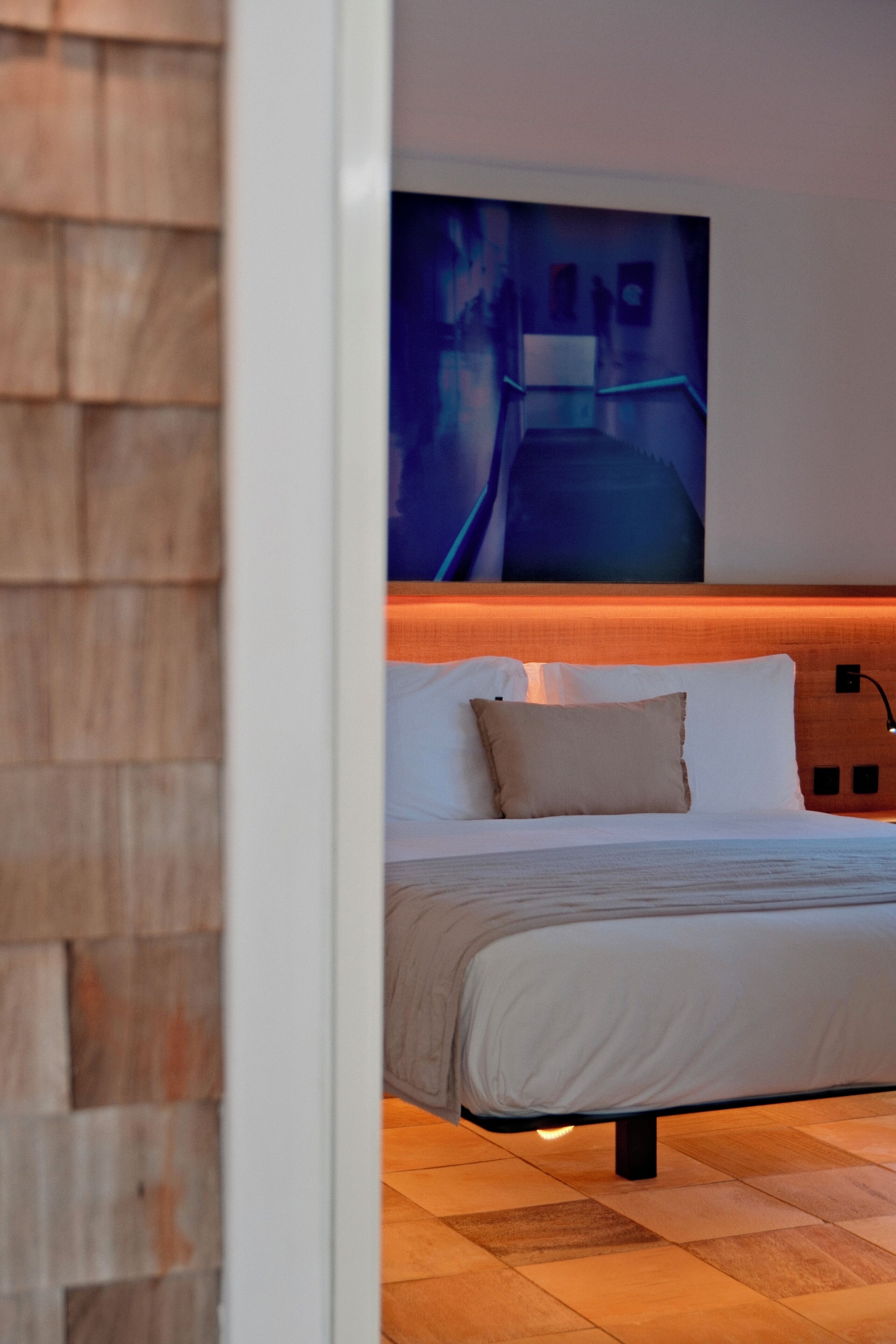 A modern hotel room with a wooden headboard, a large bed with white bedding, and a wall-mounted artwork.