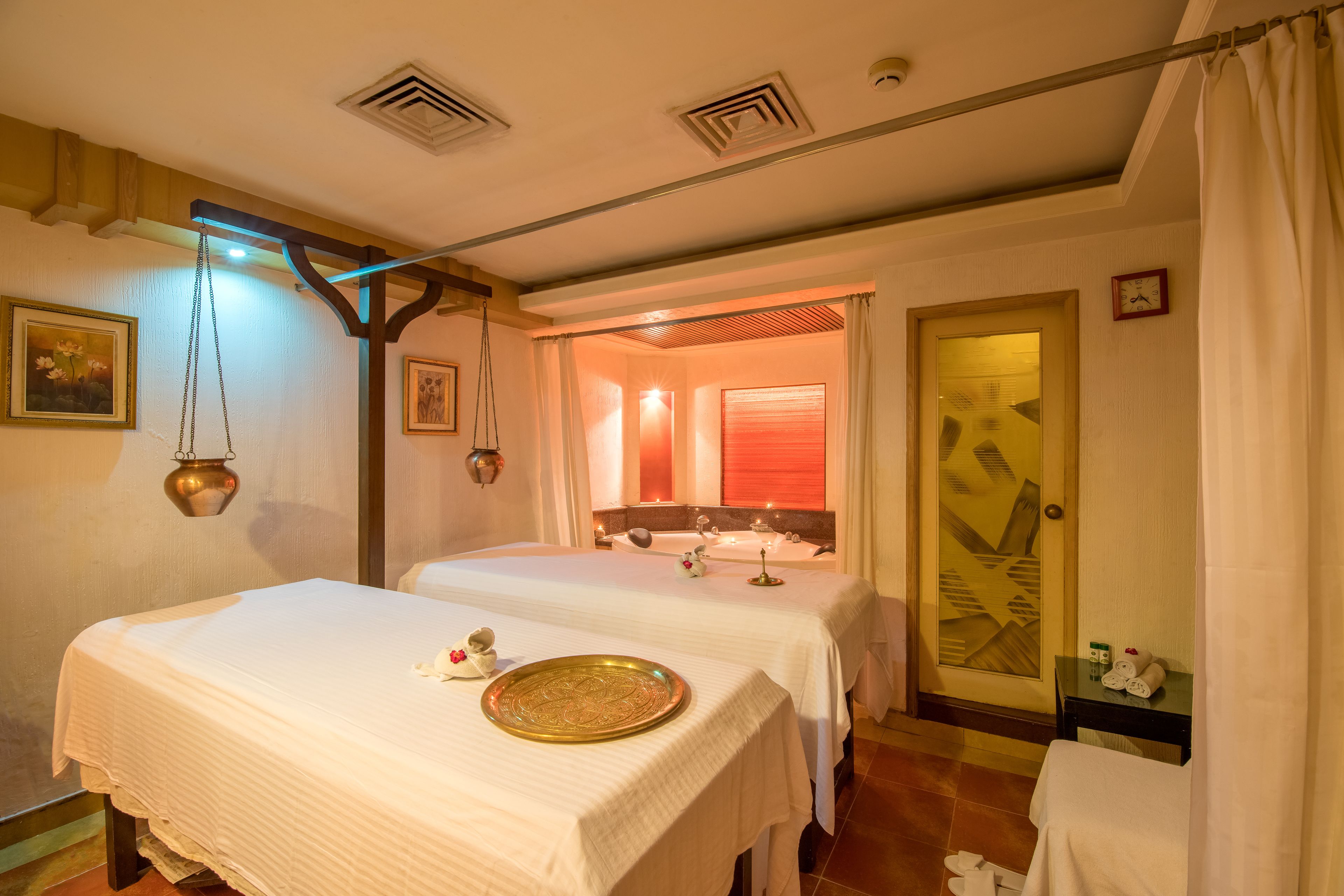Ein Wellnessraum mit zwei Betten, einer Massageliege, einem kleinen Tisch mit Handtüchern und einer Tür mit geometrischem Muster.