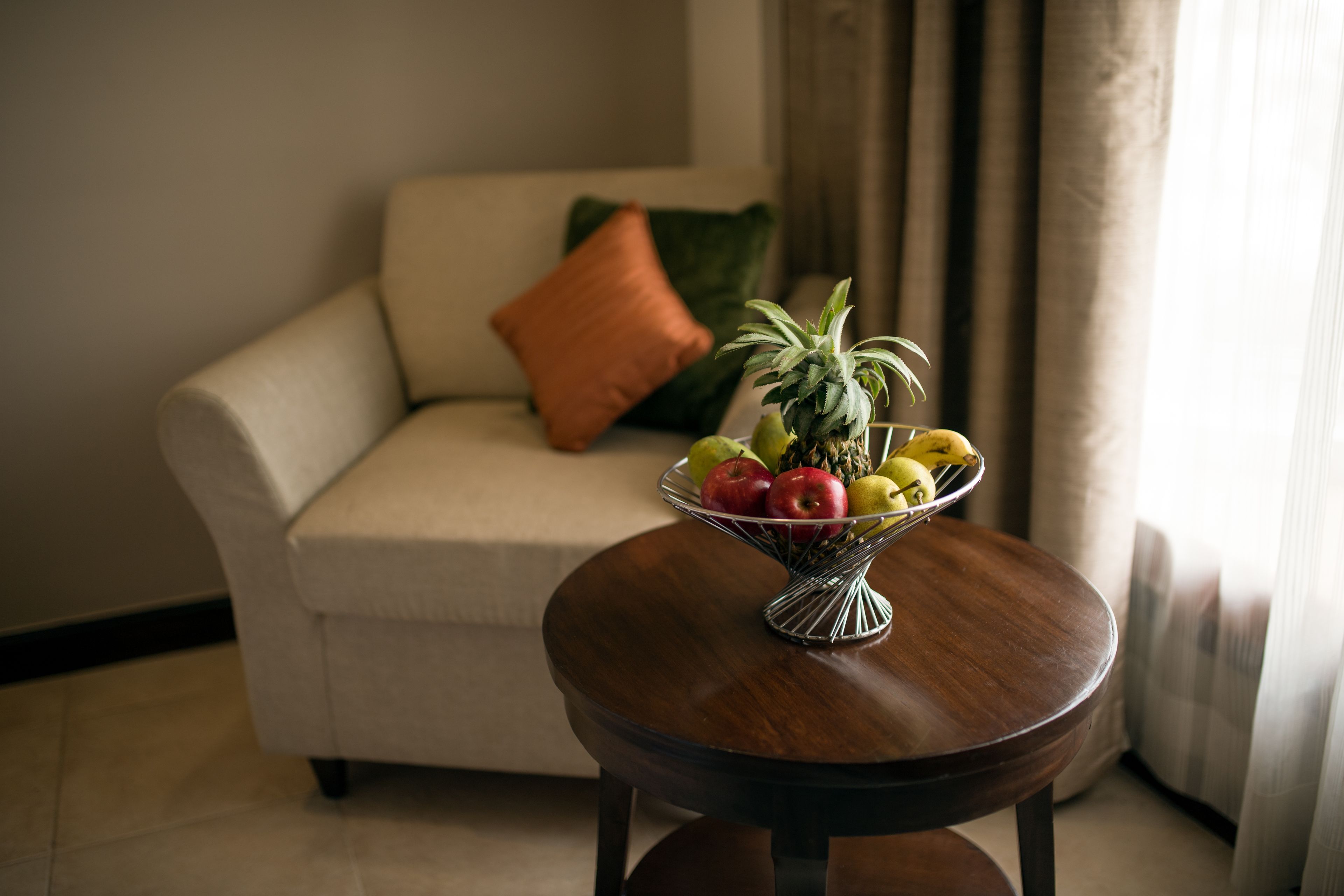 Una mesa redonda de madera con un frutero en primer plano. Un sofá color beige con un cojín naranja se ve al fondo.