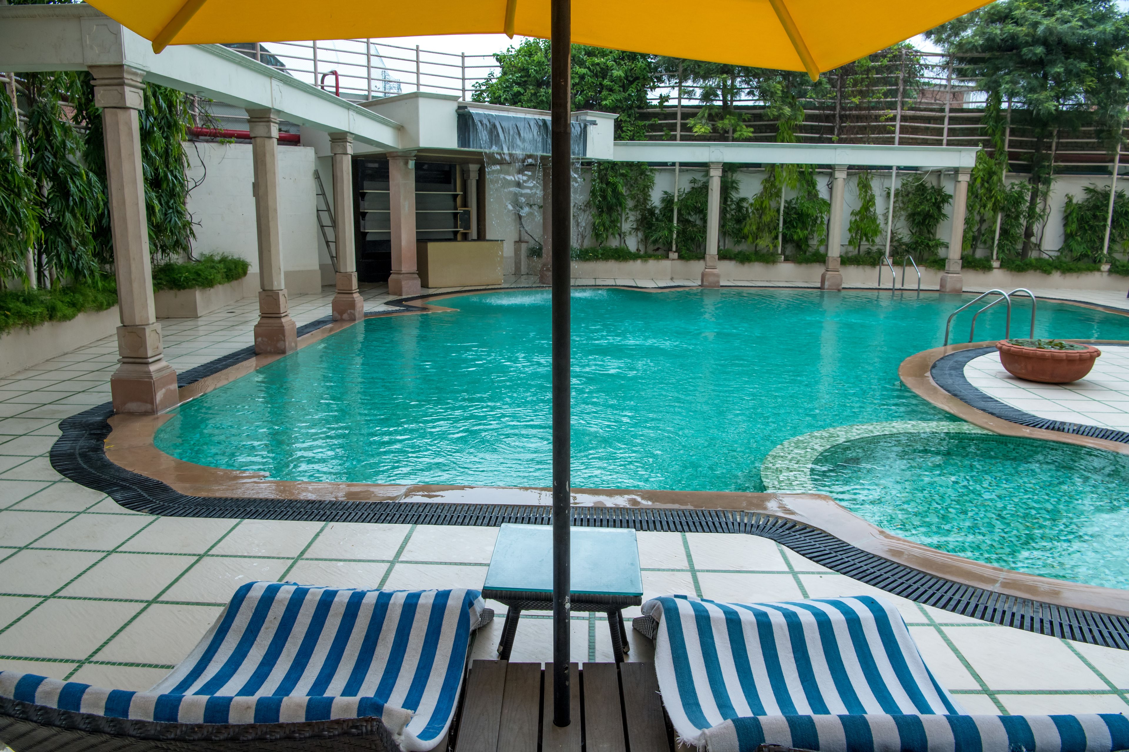 Uma piscina com uma pequena cascata, rodeada por espreguiçadeiras e uma área coberta com um guarda-sol amarelo.