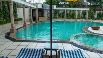 Uma piscina com uma pequena cascata, rodeada por espreguiçadeiras e uma área coberta com um guarda-sol amarelo.