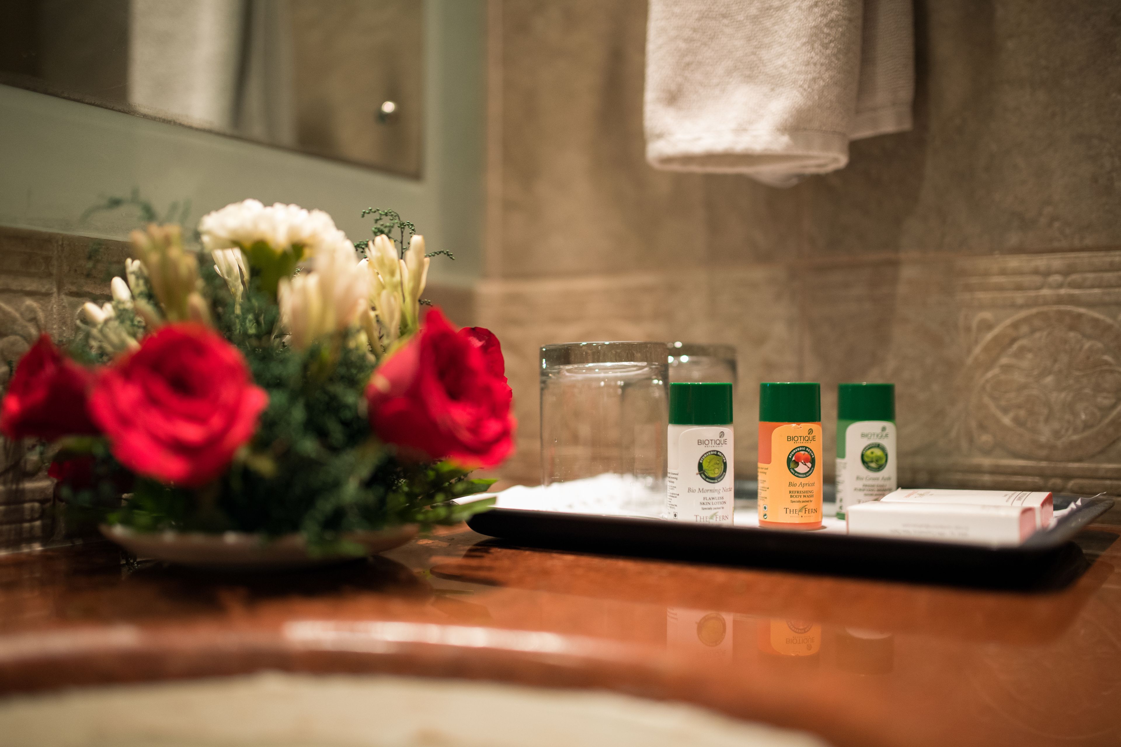 Tocador de baño con un ramo de flores rojas y blancas, un vaso de agua y dos productos para el cuidado de la piel Biotique.